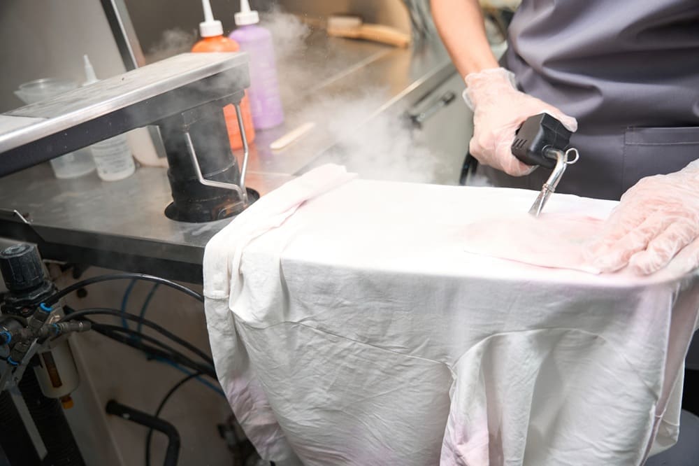 A person in gloves uses a steam cleaning tool to clean a white shirt on an industrial pressing board in a laundry facility.